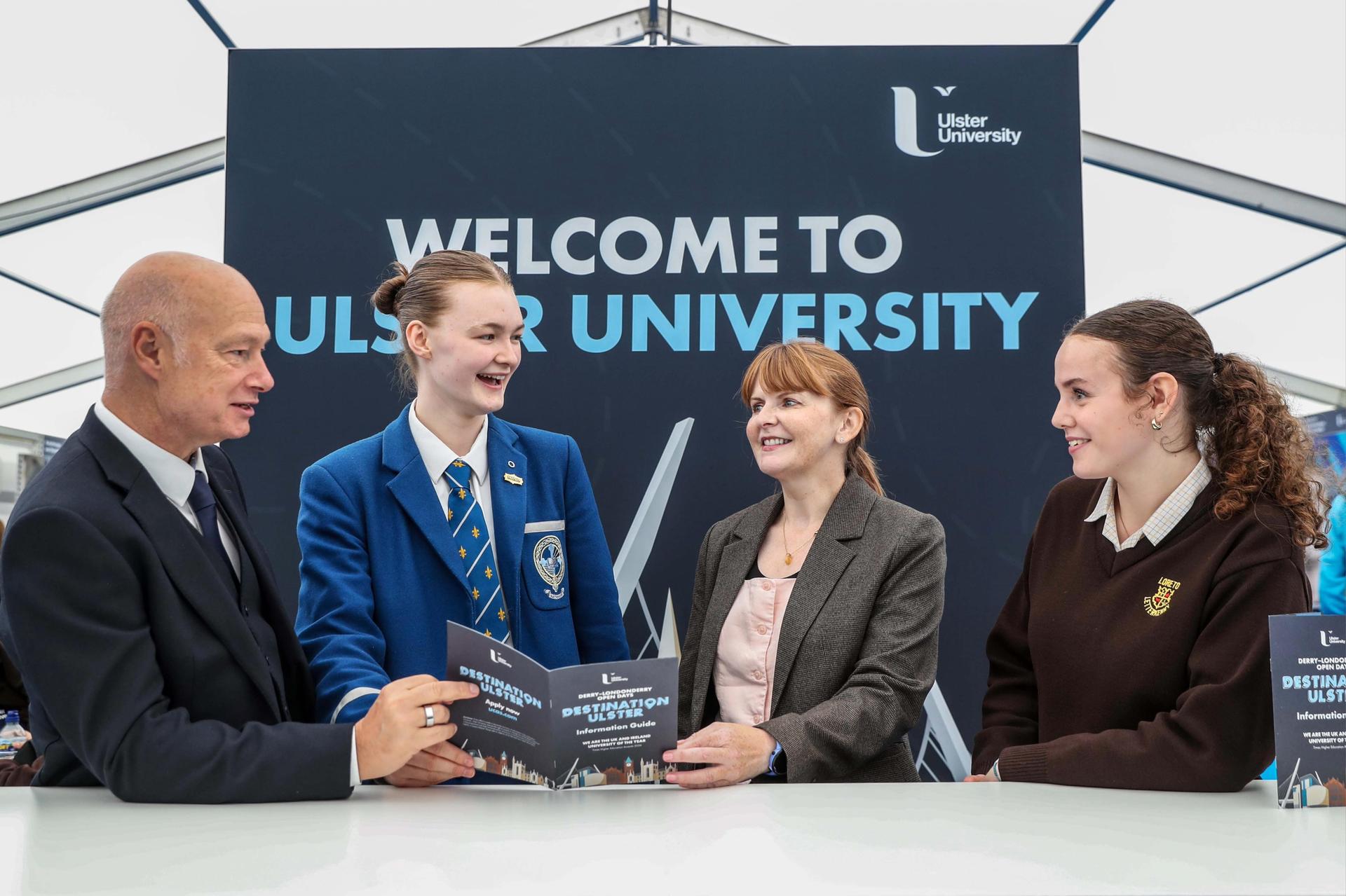 A group of four people, including two students in uniforms and two adults, sit smiling at a table with brochures. “Welcome to Ulster University” is displayed behind them.
